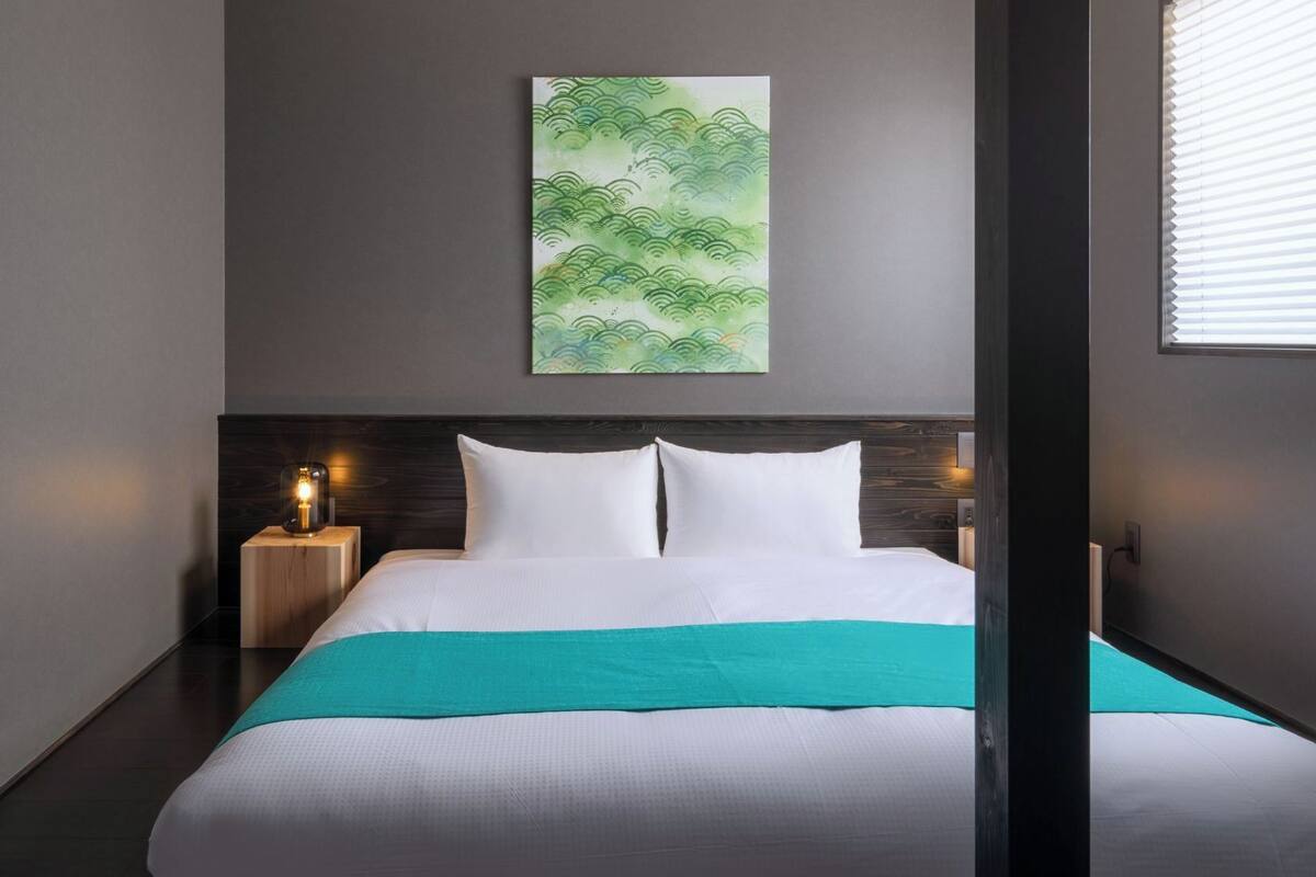 Een hotelkamer met een bed, nachtkastje, een kunstwerk aan de muur, en een raam met zonwering.