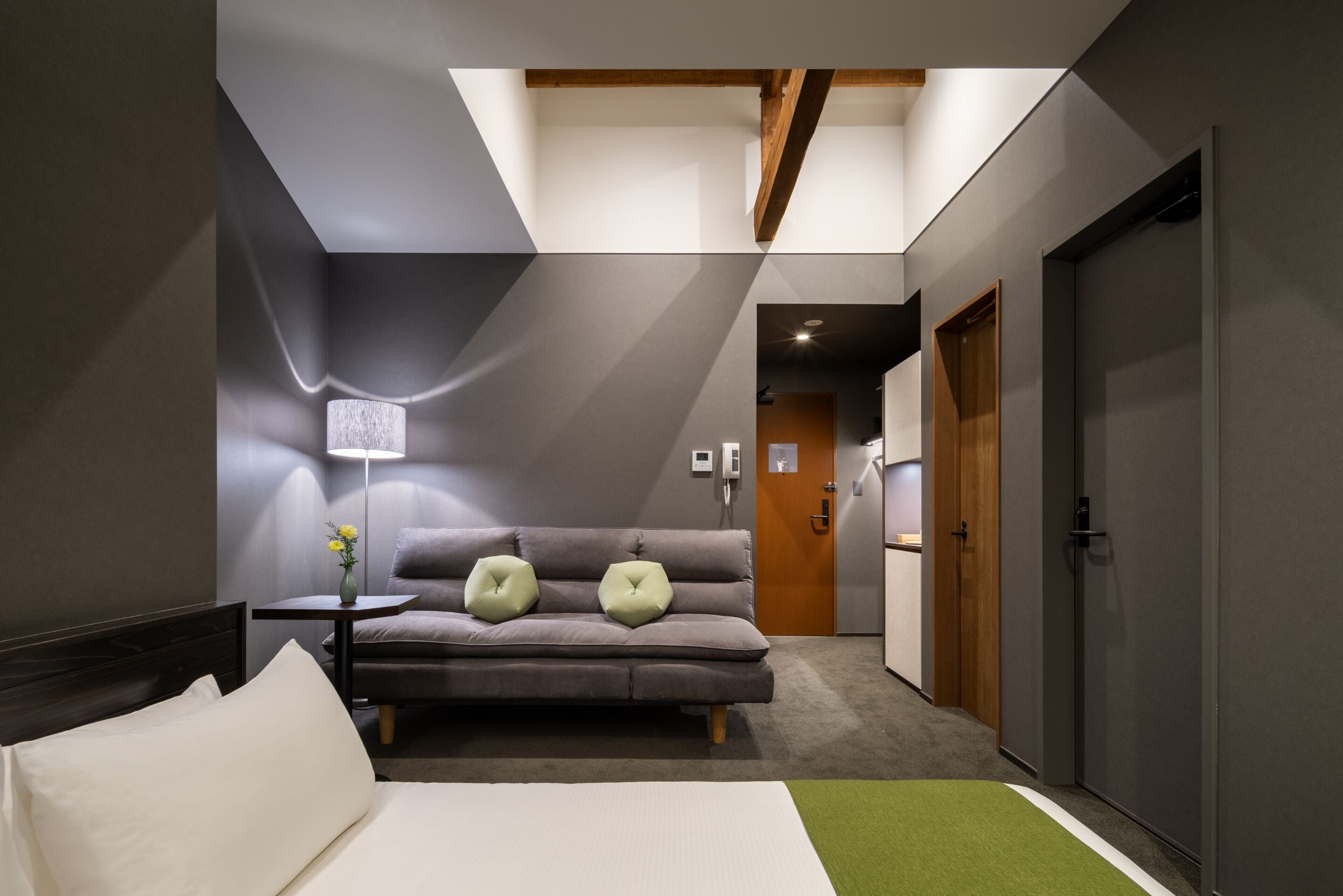 A modern hotel room with a bed, a sofa, a small table with a lamp, and a view of the corridor.