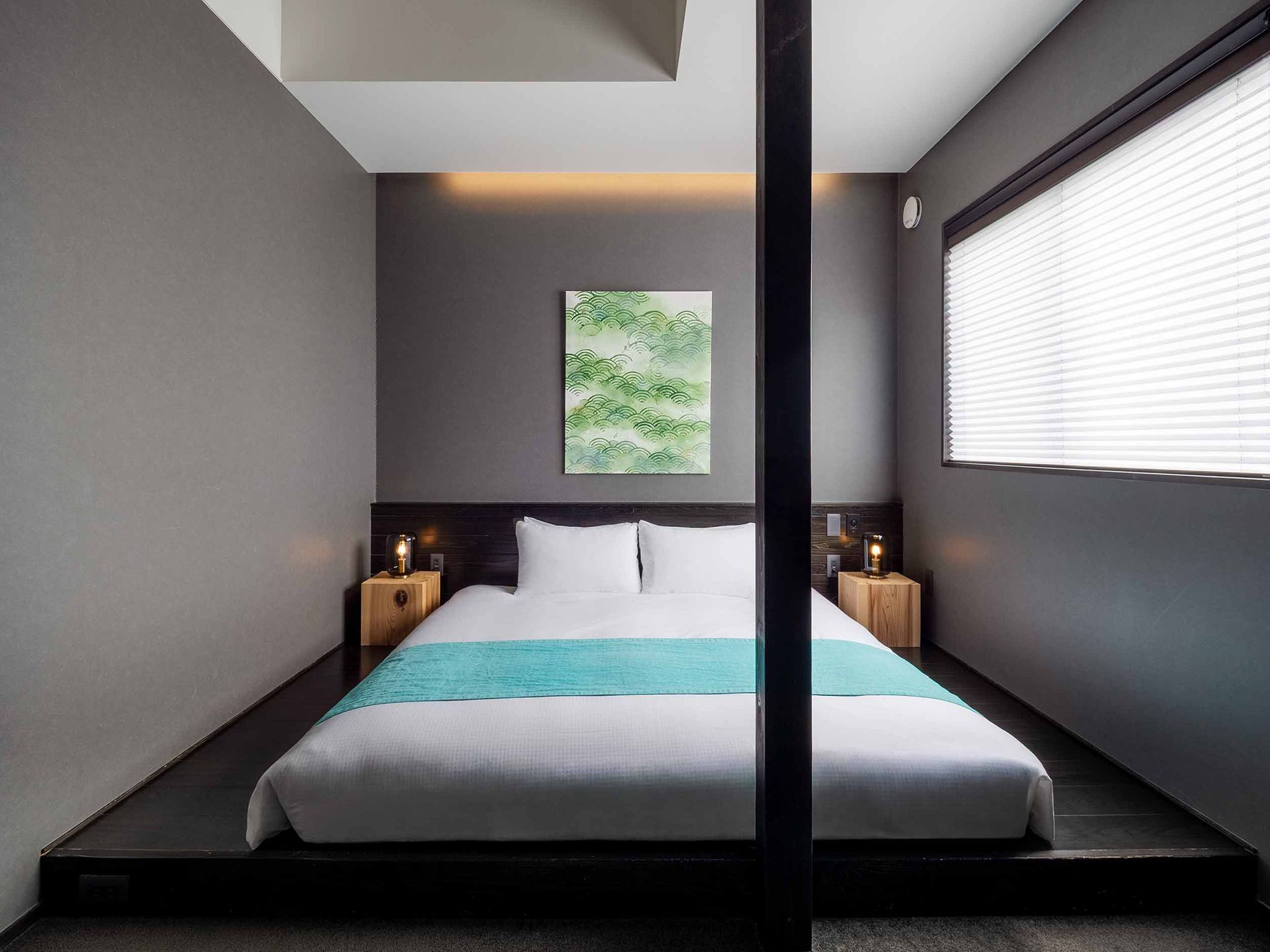 Quarto de hotel moderno com uma cama grande, duas mesas de cabeceira com luminárias, uma obra de arte na parede e uma janela com persianas.