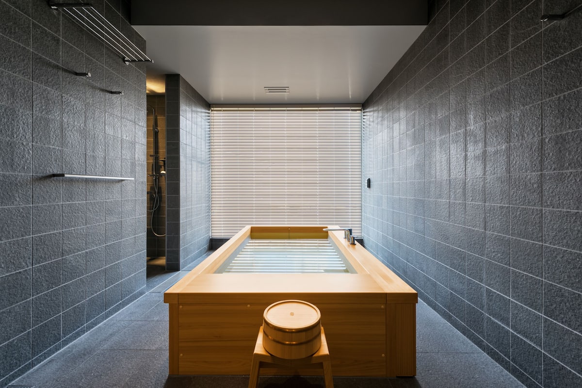 Een moderne badkamer met een houten bad, een houten krukje en een groot raam met zonwering.