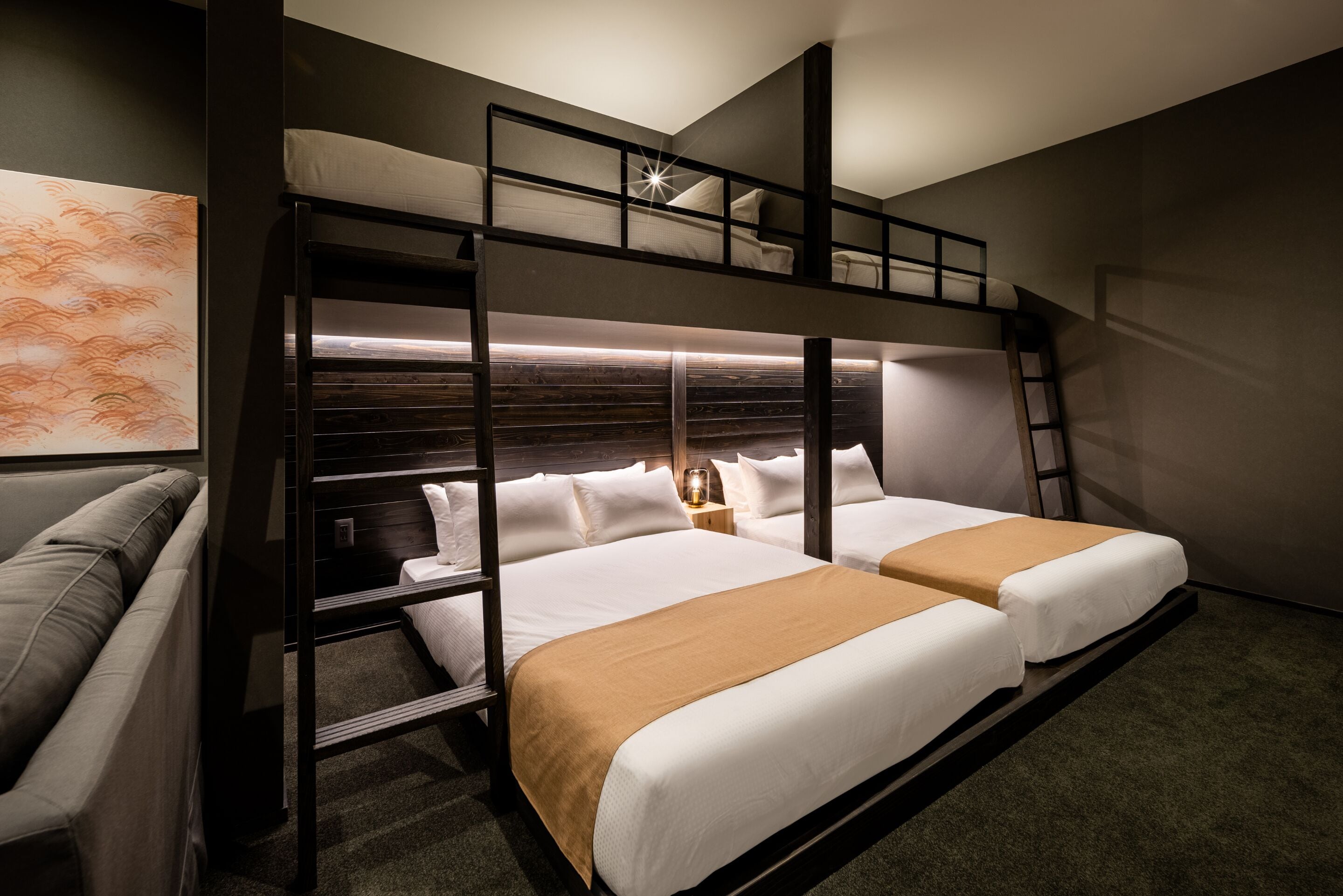 A bunk bed room with a sofa, a nightstand with a lamp, and a painting on the wall.