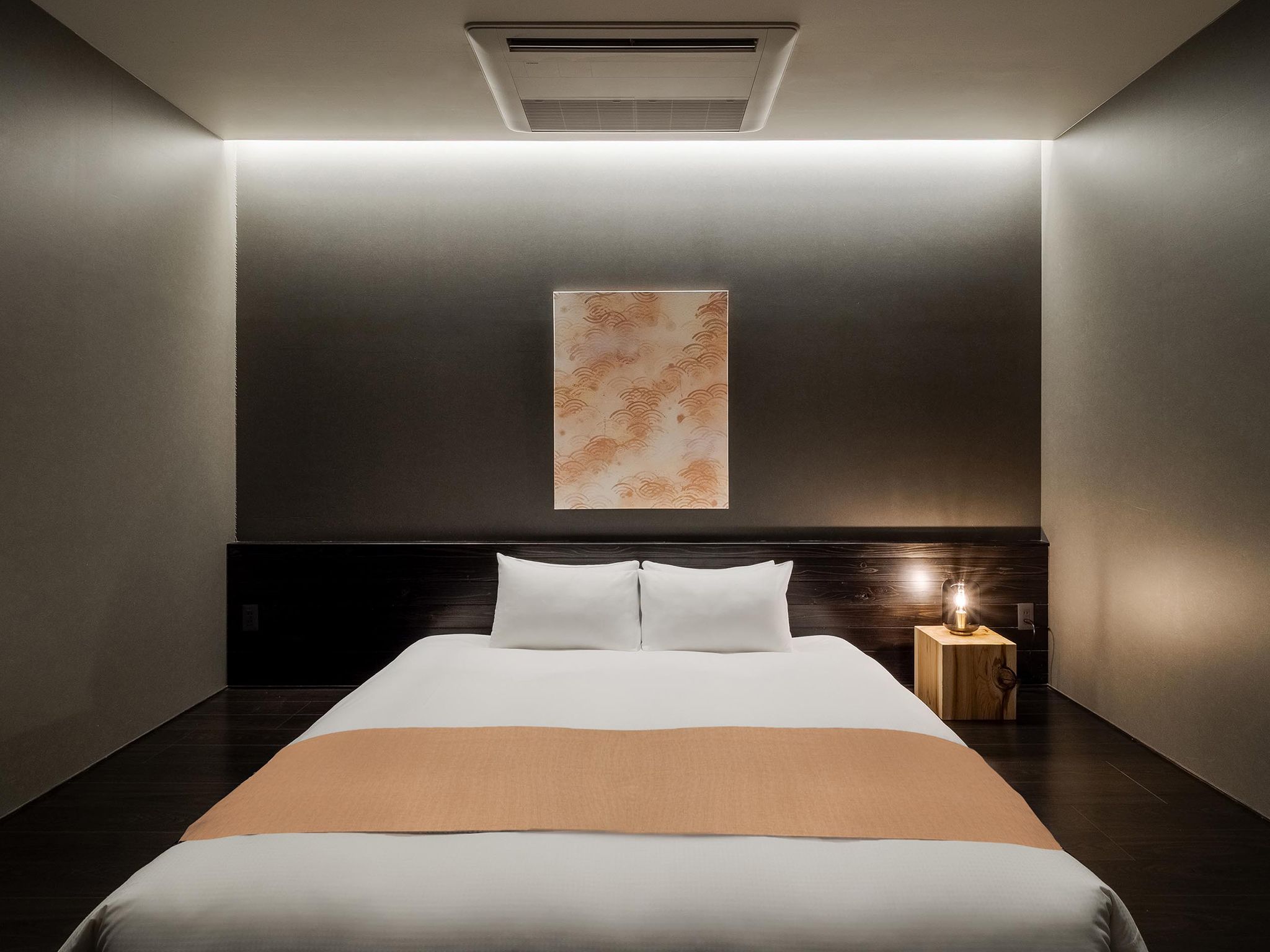 A hotel room with a bed, a nightstand with a lamp, and a wall-mounted air conditioning unit.