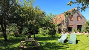 A backyard with a wooden picnic table and two lounge chairs, surrounded by trees and a brick house. - Ferienwohnung Verklicker - Ferienwohnungen im Haus am Watt in Ostbense (Neuharlingersiel)
