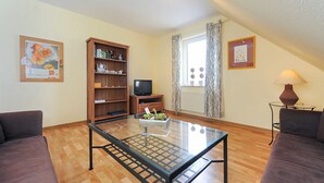 A living room with a glass coffee table, two sofas, a wooden bookshelf, a television, and a window with curtains. - Ferienwohnung Verklicker - Ferienwohnungen im Haus am Watt in Ostbense (Neuharlingersiel)