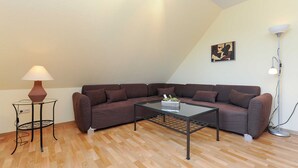 A living room with a brown sectional sofa, a glass coffee table, a floor lamp, and a painting on the wall. - Ferienwohnung Verklicker - Ferienwohnungen im Haus am Watt in Ostbense (Neuharlingersiel)