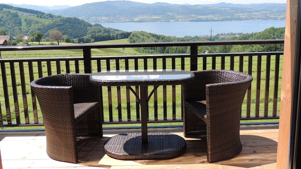 A balcony with wicker furniture, a glass table, and a view of a lake and mountains.