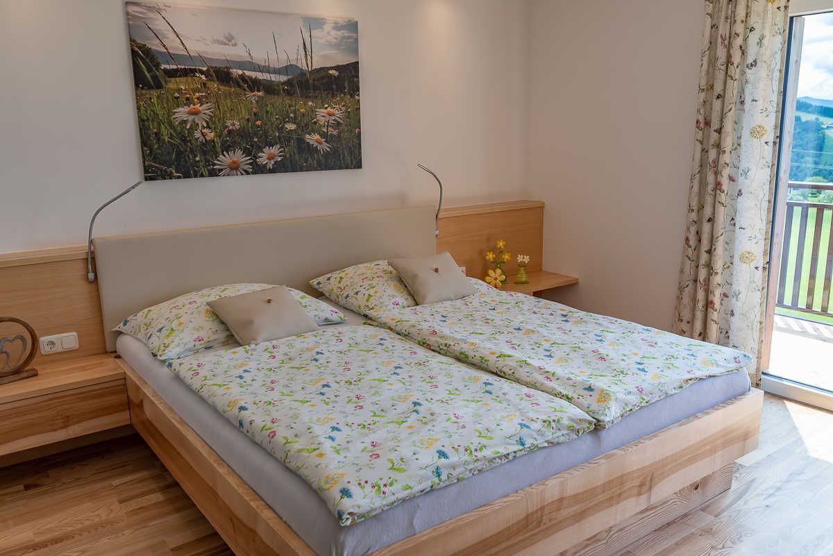 Una cama doble con ropa de cama floral, una mesita de noche con lámpara, un cuadro colgado en la pared y una ventana con cortinas.
