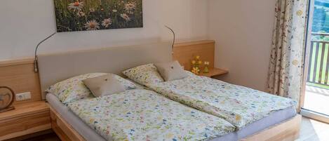 Una cama doble con ropa de cama floral, una mesita de noche con lámpara, un cuadro colgado en la pared y una ventana con cortinas.