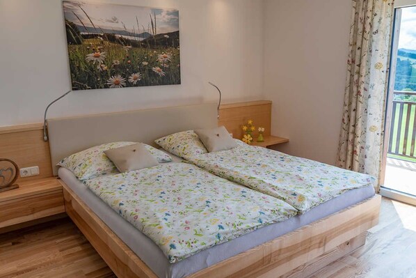 A double bed with floral bedding, a bedside table with a lamp, a wall-mounted painting, and a window with curtains.