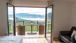 A room with a bed, a sofa, a balcony with wicker chairs, and a table with flowers.