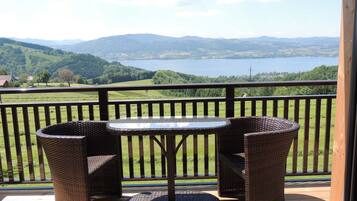 Un balcon agrémenté de meubles en osier, d’une table en verre et offrant une vue sur un lac et des montagnes.