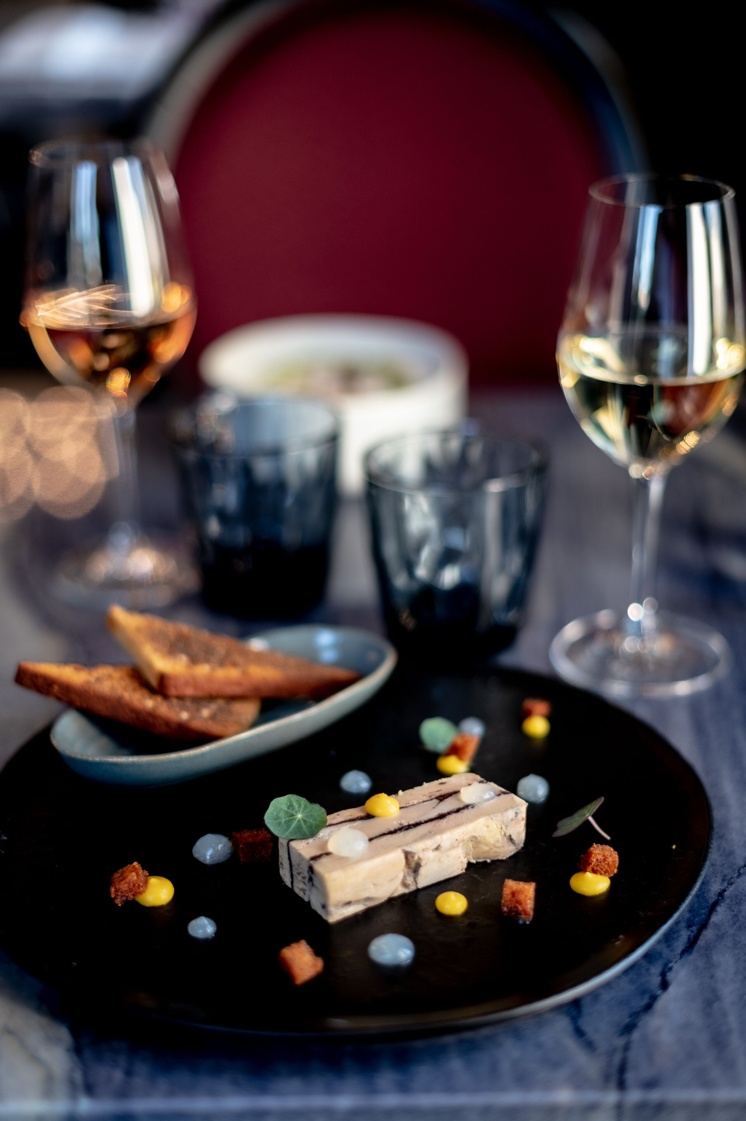 Un plato de postre con una porción de pastel, adornos y dos copas de vino sobre una mesa.