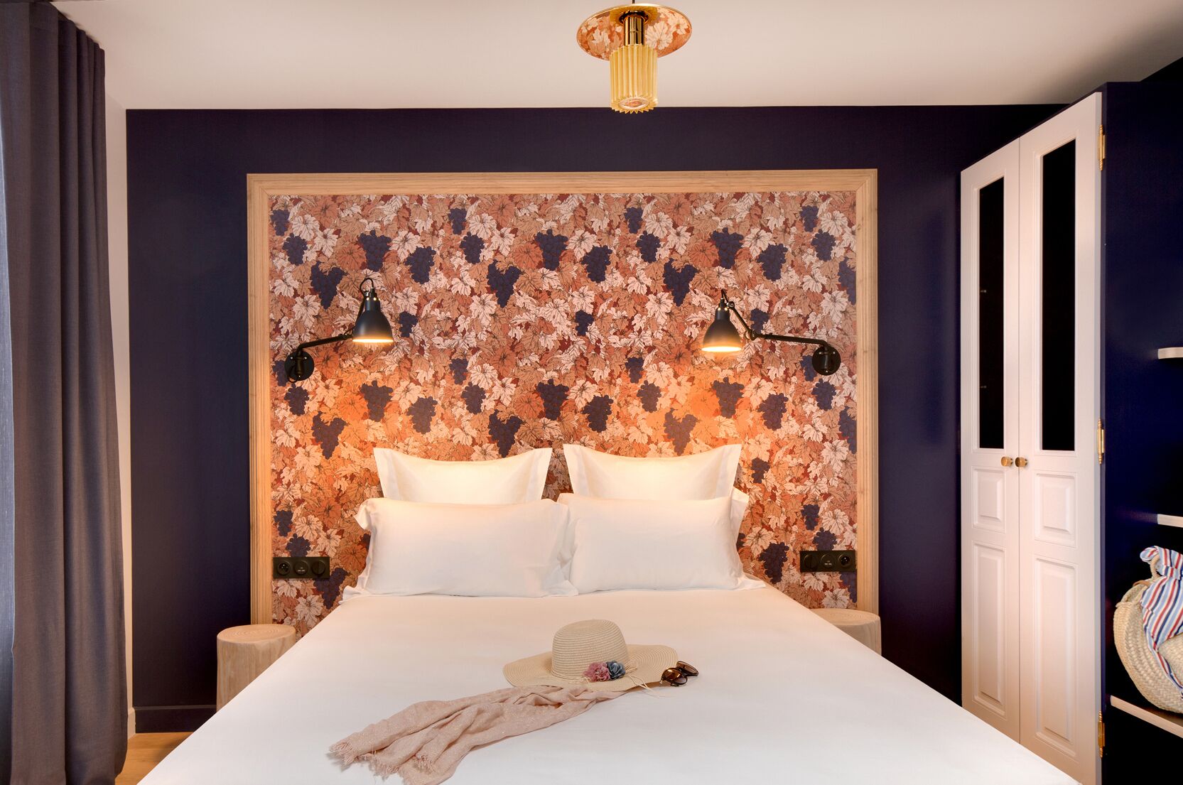 Une chambre d’hôtel avec un grand lit, deux lampes de chevet, une tête de lit à motifs floraux et une armoire sur laquelle est posé un sac à rayures.