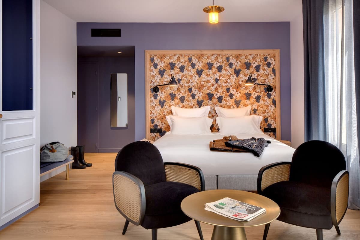 A hotel room with a large bed, two armchairs, a small round table with a newspaper, and a floral-patterned headboard.