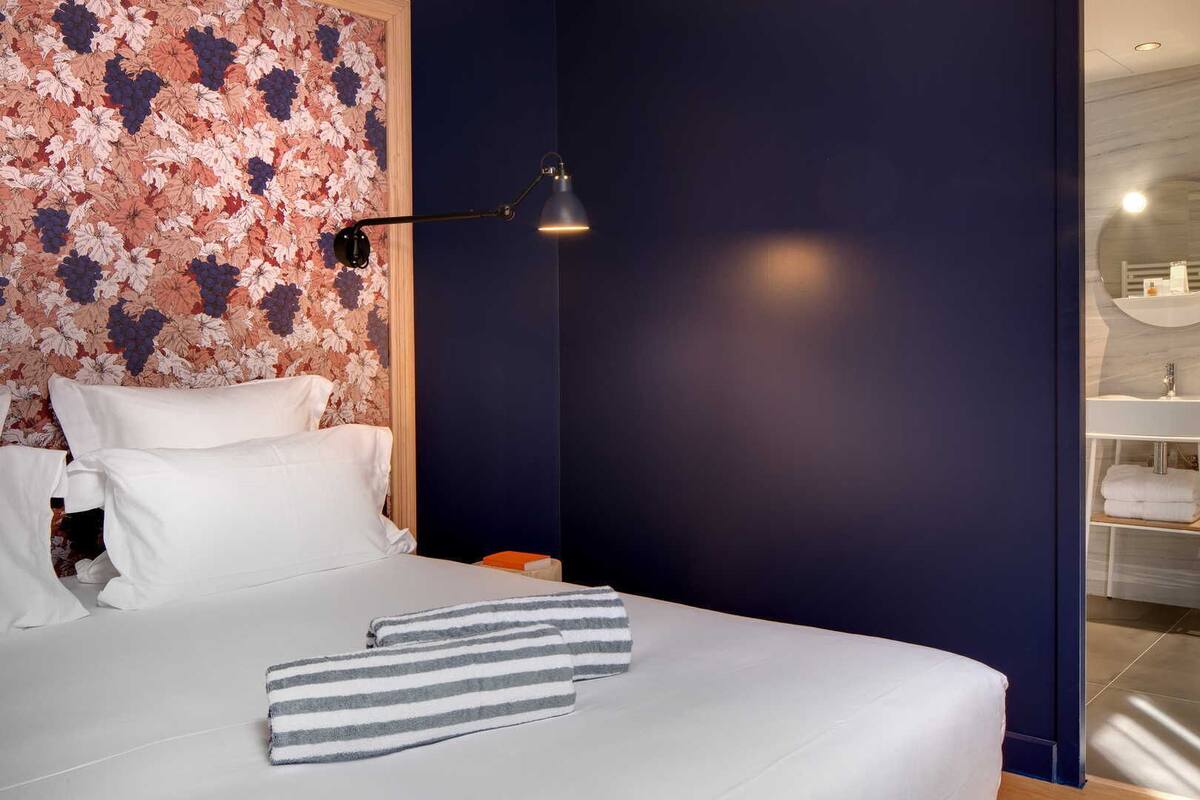 A hotel room with a large bed, a nightstand, a desk, a desk lamp, and a bathroom with two sinks and a mirror.