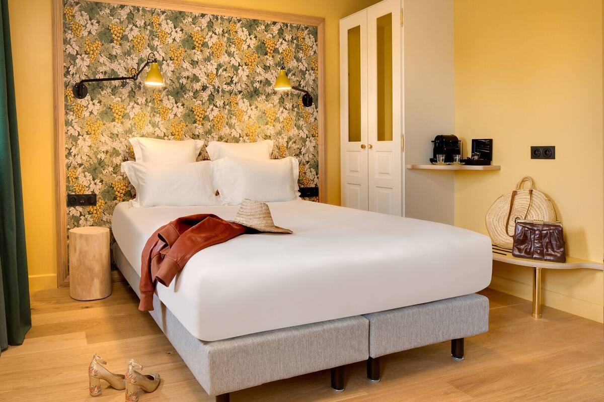 A neatly made bed with pillows, a bedside table with a lamp, a wardrobe, and a small shelf with items.