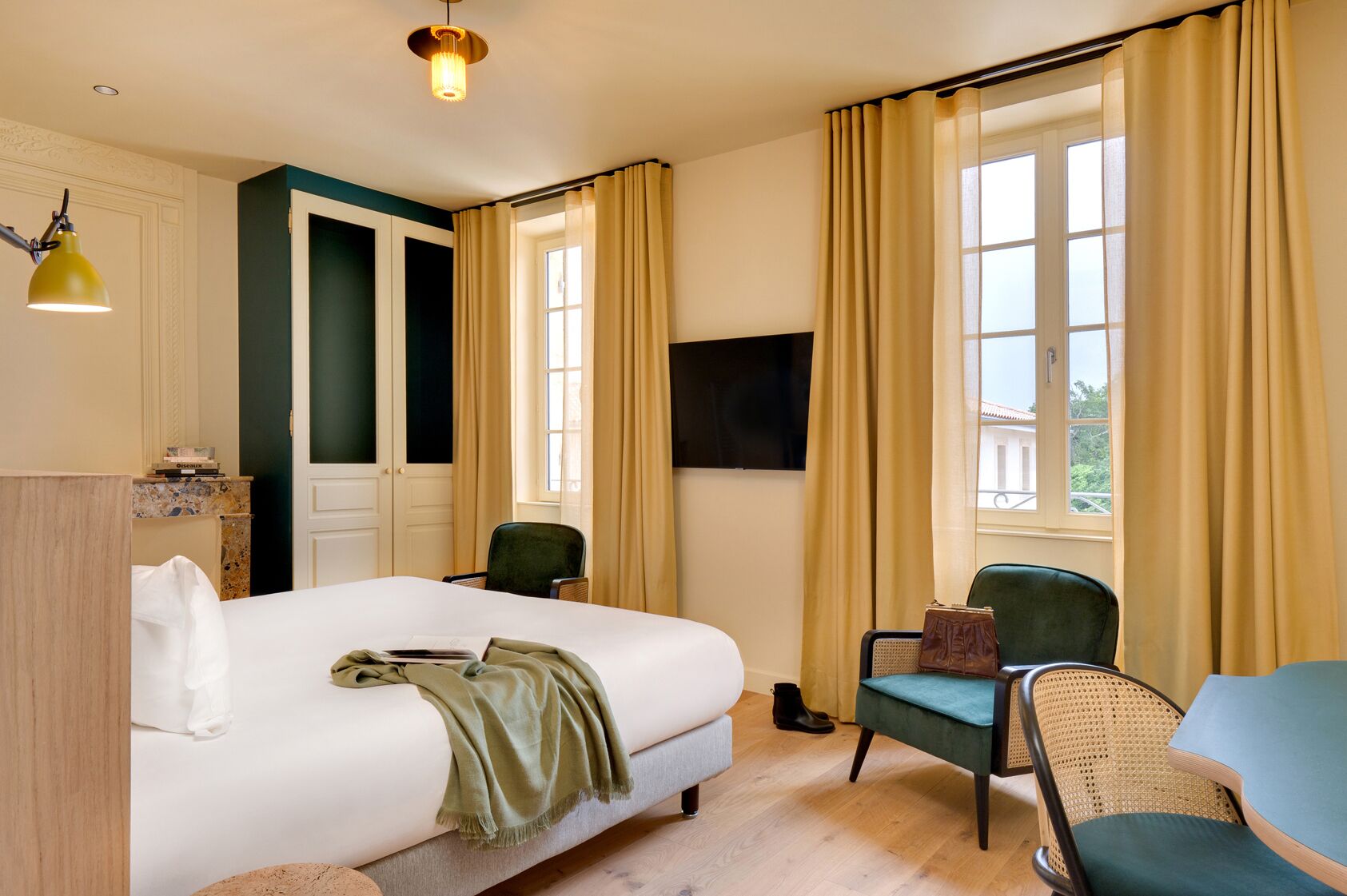 Habitación de hotel con cama, dos sillas, escritorio, televisor y ventanas amplias con cortinas.