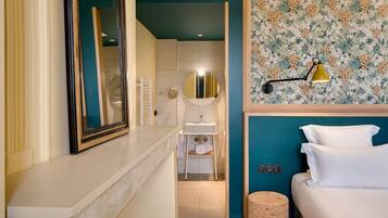 A hotel room with a white vanity, a mirror, a sink, a toilet, and a bed with white pillows.