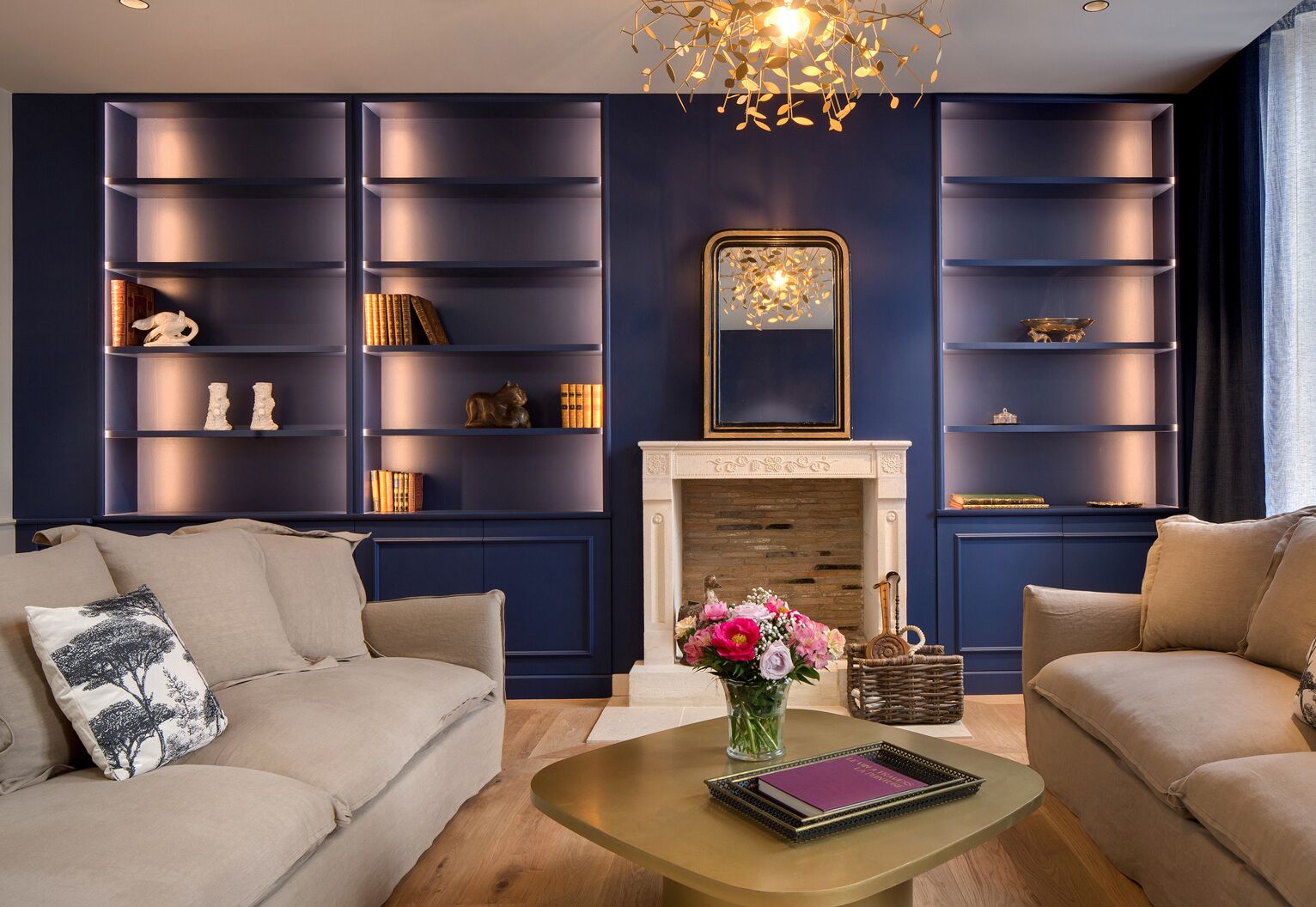 A modern living room with a dark blue wall, built-in shelving, a fireplace, and a coffee table with a book and flowers.