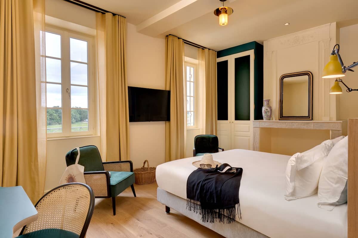 A hotel room with a bed, two chairs, a television, and a window with curtains.