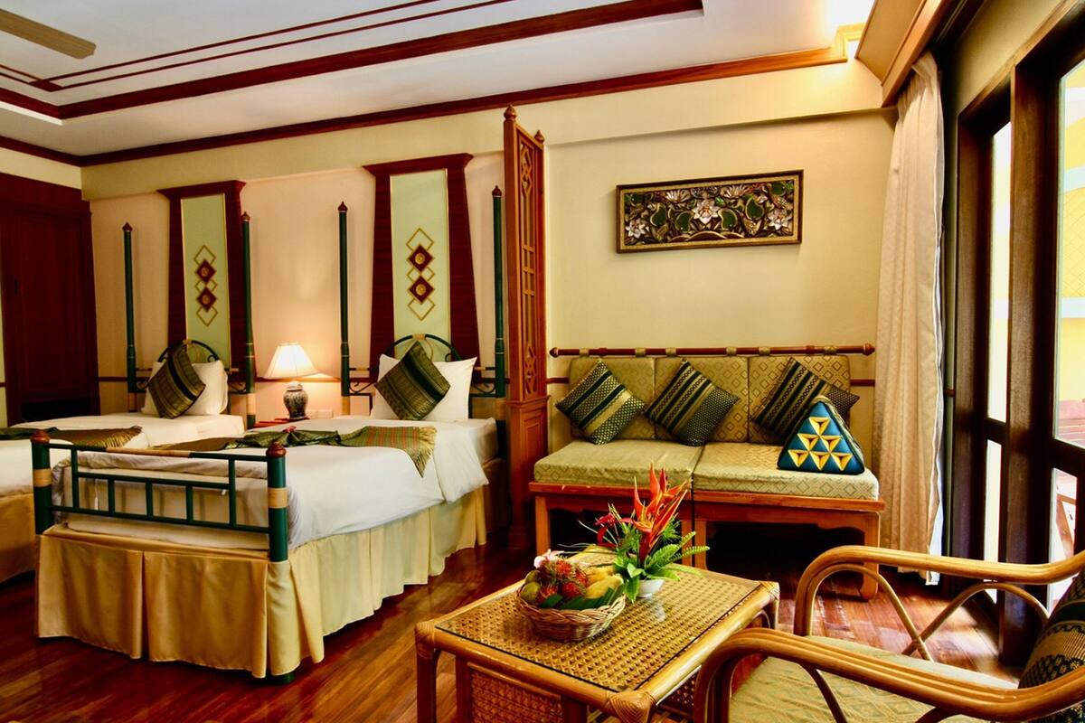 A hotel room with two beds, a sofa, a table with a fruit basket, and a window with curtains.
