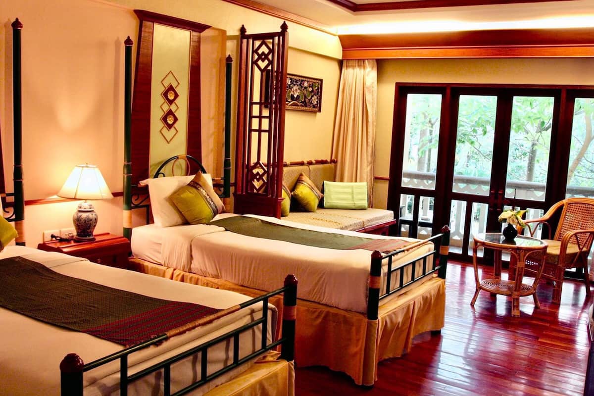 A hotel room with two beds, a sitting area with a chair, a table, and a view of greenery through large windows.
