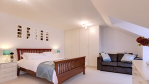 A bedroom with a wooden bed, a nightstand with a lamp, a dresser, a sofa, and a radiator. - BookedUK - Penthouse 3 Bedroom Apartment (Bishop's Stortford)