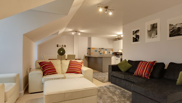 A modern living room with a sofa, ottoman, and a dark-colored couch. There is a kitchen area in the background with a tiled backsplash and a bar counter. - BookedUK - Penthouse 3 Bedroom Apartment (Bishop's Stortford)