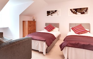 Two single beds with white duvets and striped pillows, a dresser, and a lamp, set against a wall with framed artworks and a grey armchair.