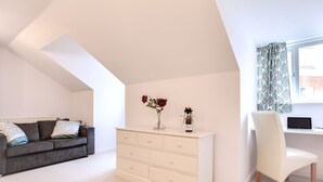 A modern bedroom with a loft-style ceiling, a white dresser, a grey sofa, and a white chair. - BookedUK - Penthouse 3 Bedroom Apartment (Bishop's Stortford)