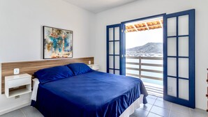 Ein Schlafzimmer mit einem Bett, einem Nachttisch, einem Gemälde und Blick durch ein blau gerahmtes Fenster.