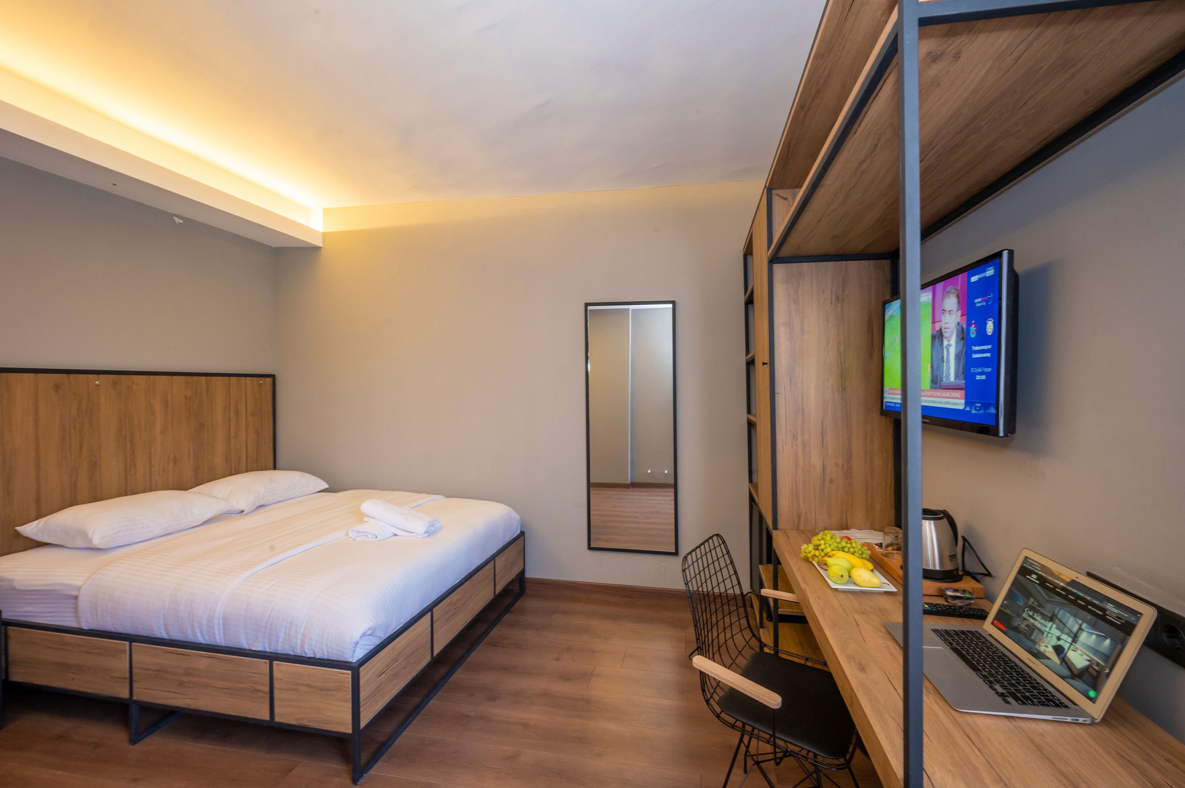 Hotellihuone, jossa on sänky, työpöytä kannettavalla tietokoneella, tuoli, televisio ja peili.