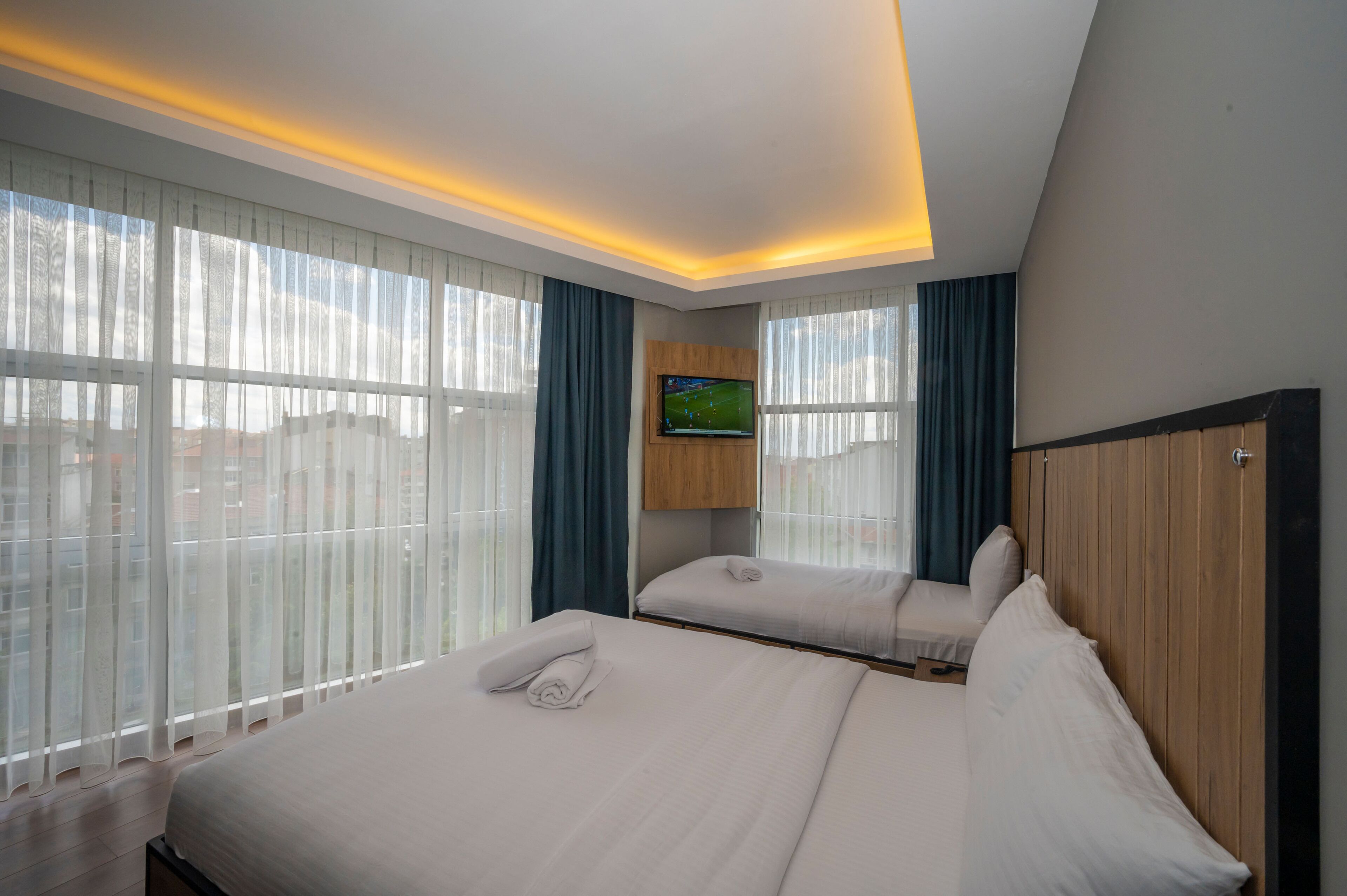 A hotel room with two beds, a flat-screen TV mounted on the wall, and large windows with sheer curtains.