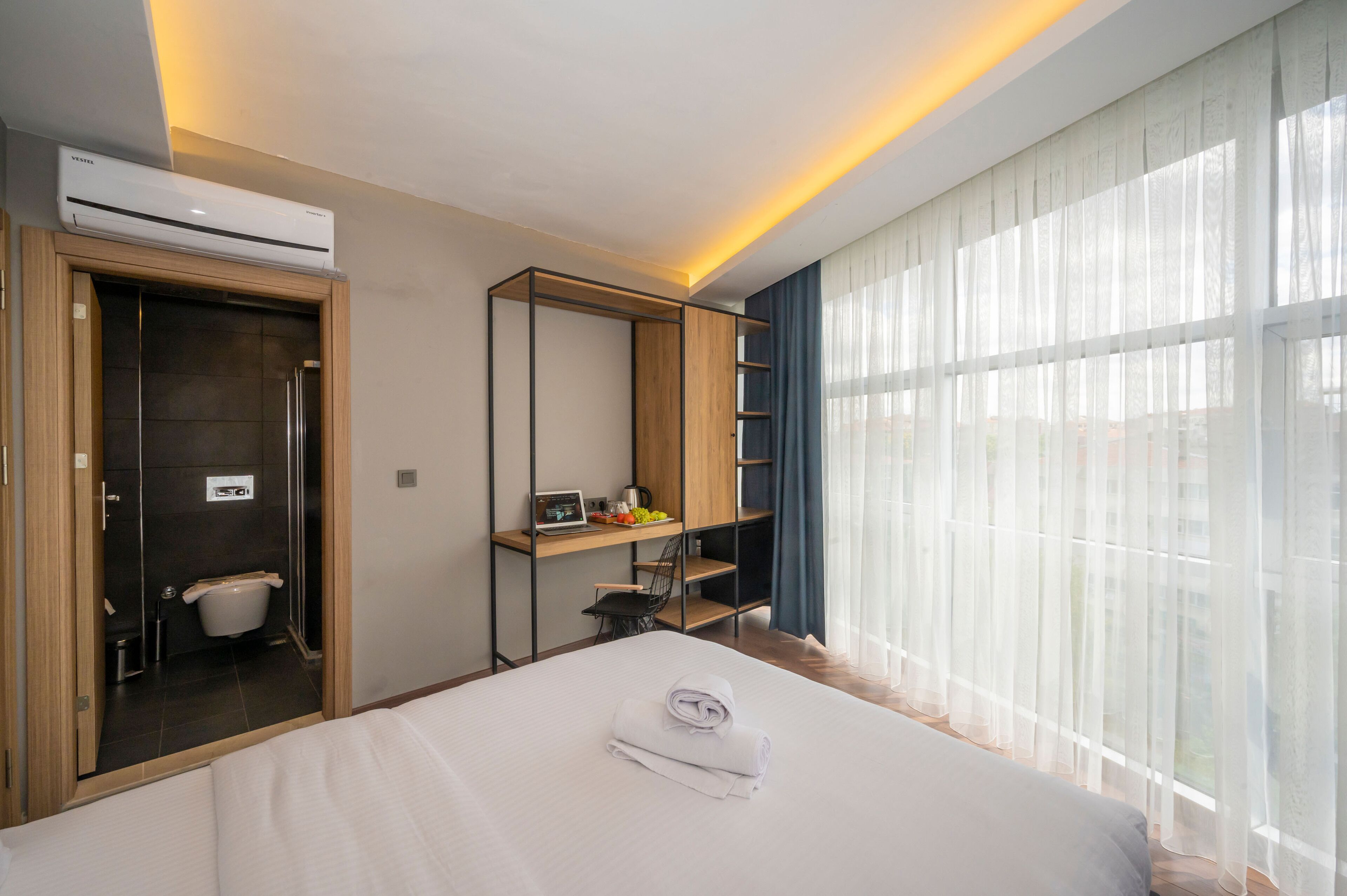 Una habitación de hotel con cama, un escritorio con computadora, un baño pequeño y un ventanal con cortinas traslúcidas.