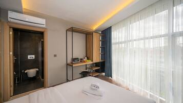 Una habitación de hotel con cama, un escritorio con computadora, un baño pequeño y un ventanal con cortinas traslúcidas.