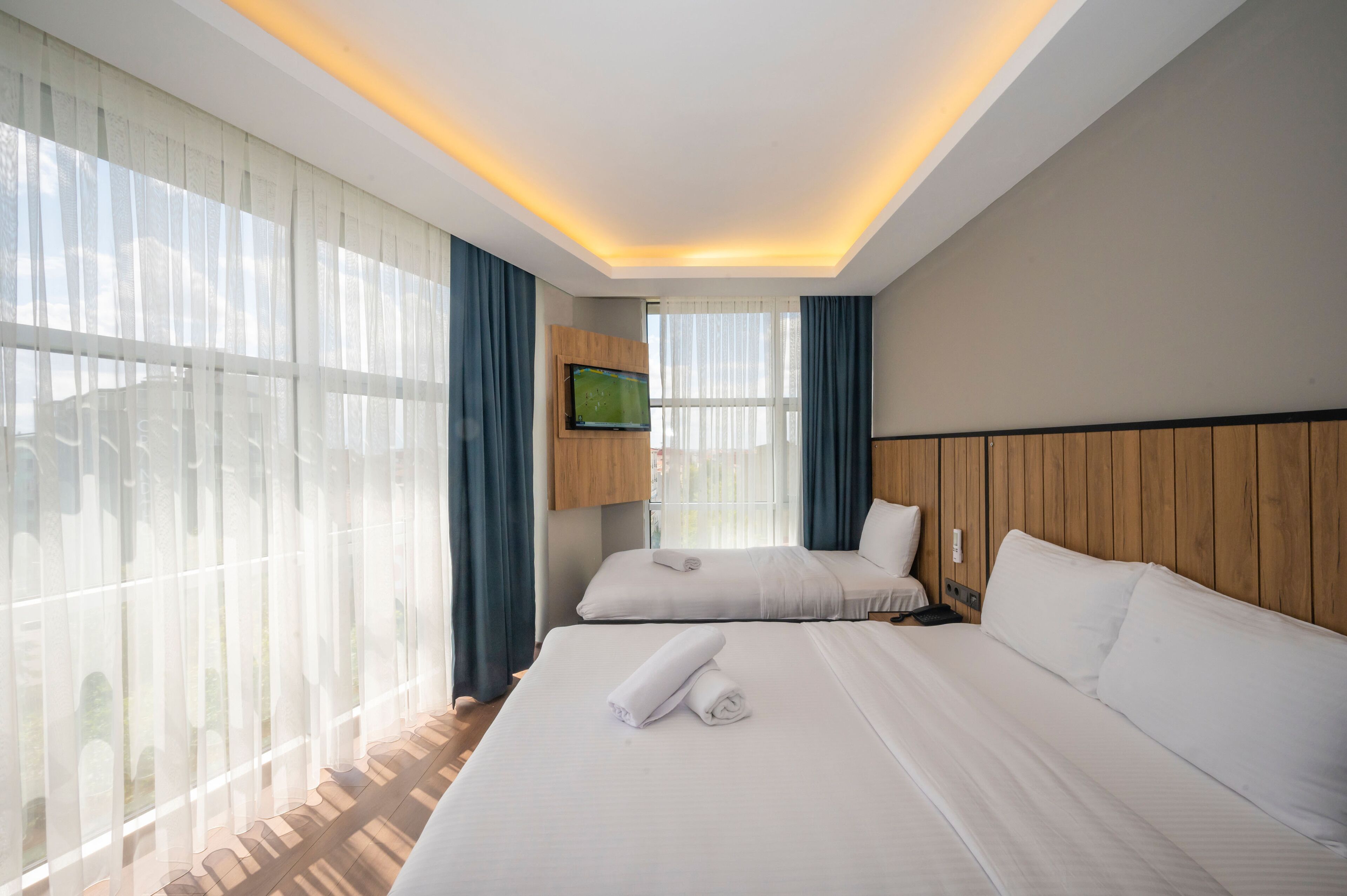 A hotel room with two beds, a flat-screen TV, and large windows with sheer curtains.