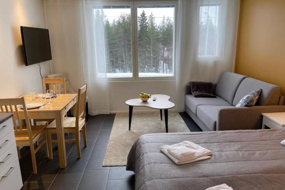 Habitación de hotel con cama, sofá, mesa de comedor con sillas, una mesita redonda con un bol y control remoto, y un televisor montado en la pared.