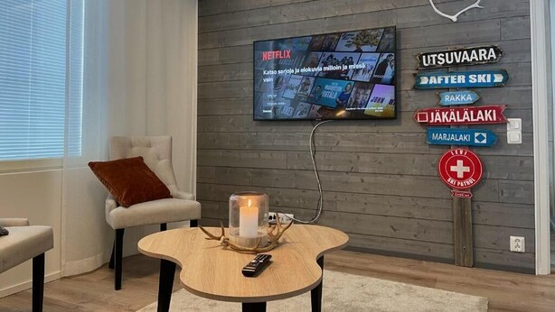 Una sala de estar moderna con un televisor montado que muestra contenido de Netflix, un rincón de asientos acogedor con una silla y una mesita, y un letrero con flechas indicativas.