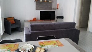 Ein modernes Wohnzimmer mit einem grauen Sofa, einem Esstisch mit orangefarbenen Platzsets, einem Fernseher und einem Kamin.