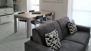Ein modernes Wohnzimmer mit einem grauen Sofa, zwei gemusterten Kissen, einem Esstisch mit Stühlen, einer Küche im Hintergrund und einem gerahmten Bild an der Wand.