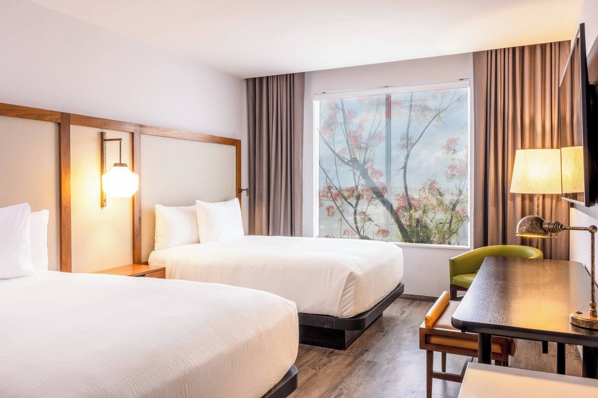 Hotellihuone, jossa on kaksi sänkyä, työpöytä, tuoli ja suuri ikkuna, josta avautuu näkymä kirsikankukkiin.