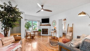 Ein geräumiges Wohnzimmer mit Holzböden, einem Kamin, einem Deckenventilator und einem großen Fenster.