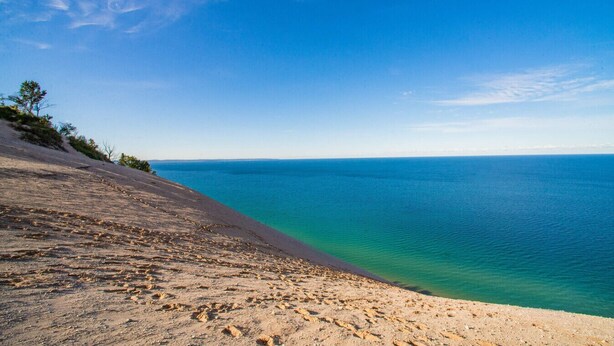 Ein Strand mit klarem, blauem Wasser, sandigem Ufer und spärlicher Vegetation.