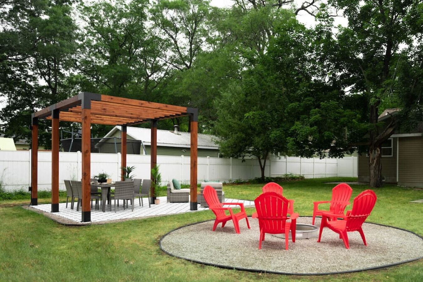 Een achtertuin met een houten pergola, een eettafel met stoelen en een vuurkorf omringd door rode Adirondack-stoelen.