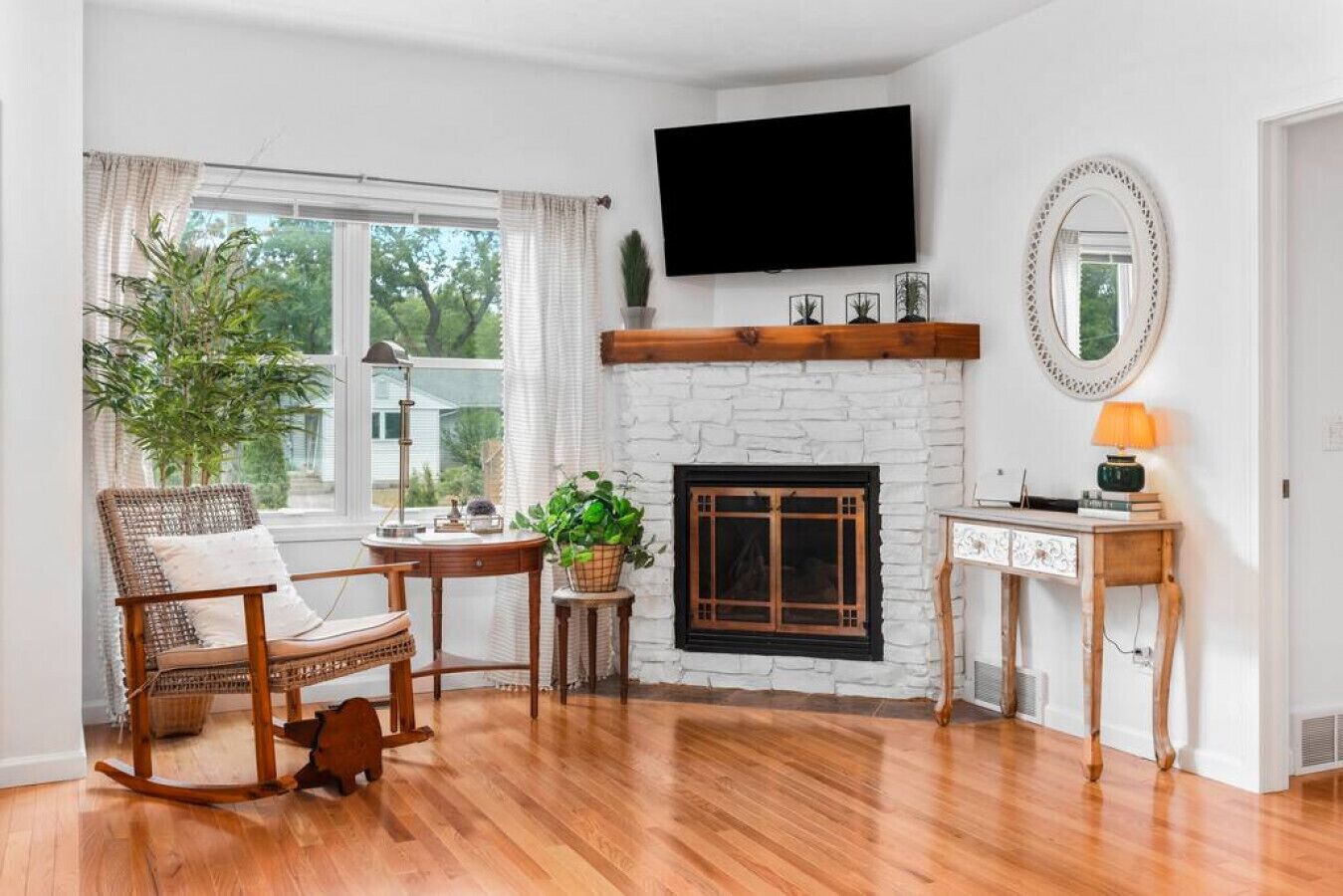 Una sala de estar con chimenea, un televisor montado encima, un sillón de mimbre, una pequeña mesa con una planta y una mesita de noche con una lámpara.