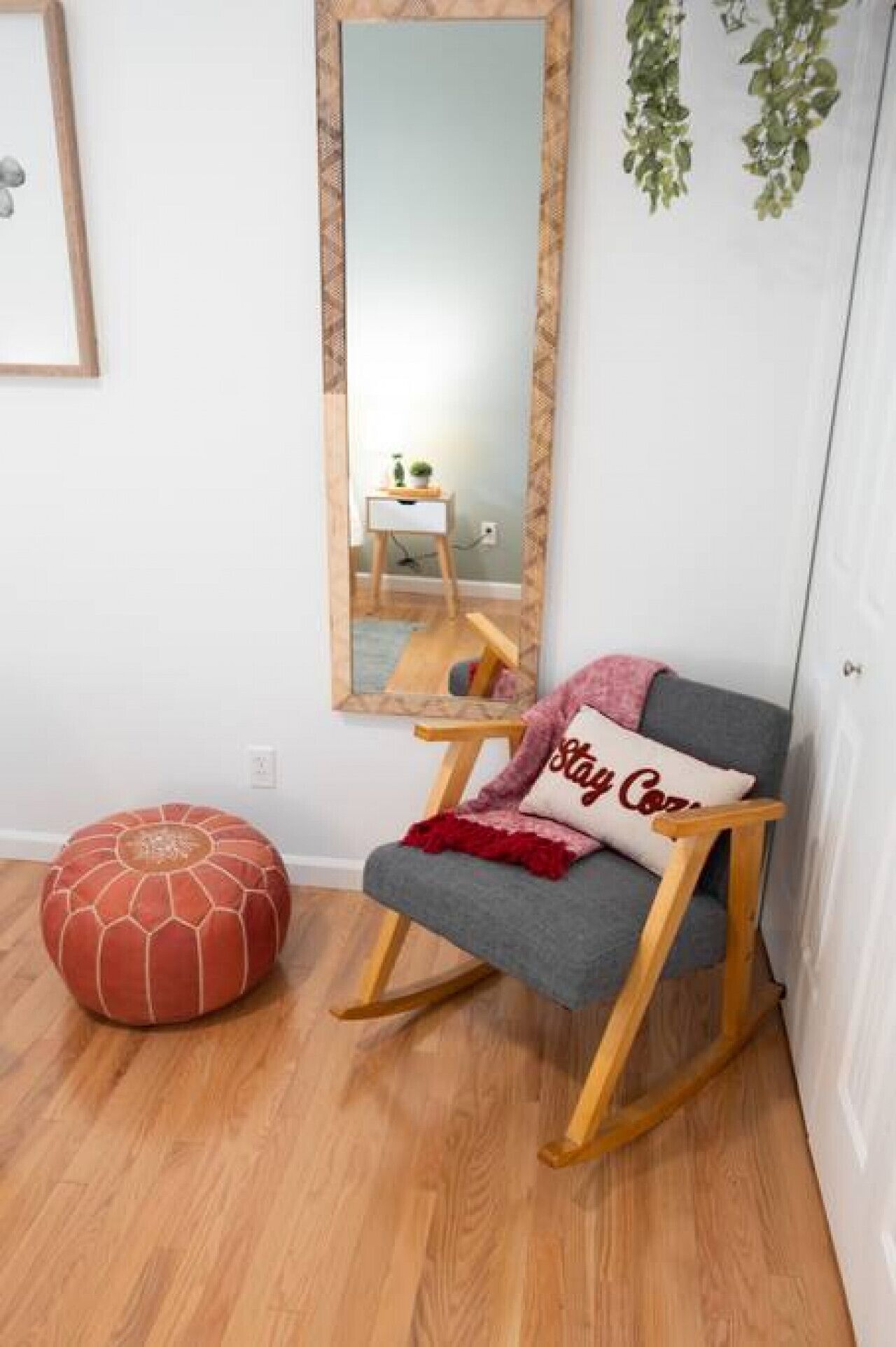 A room with a wooden rocking chair, a red pouf, a mirror, and a small table with a plant.