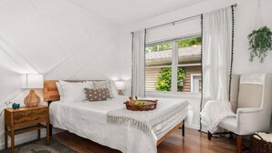 Quarto com cama, mesa de cabeceira, cadeira e janela com cortinas.