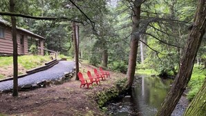 A cabin with a gravel path leading to red chairs by a stream.