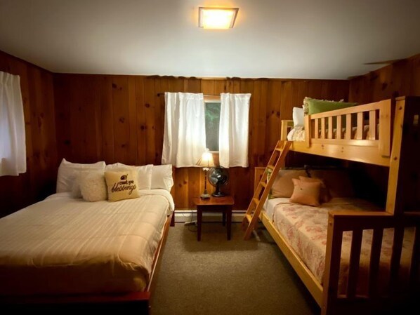 A room with two beds, one on the left and one on the right, a nightstand with a lamp, and a window with curtains.