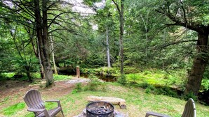 A forested area with a fire pit and two Adirondack chairs.